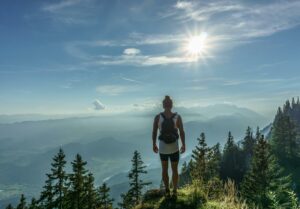 A lone hiker stands on a mountain top, overlooking a breathtaking landscape in Slovenia under the bright sun.