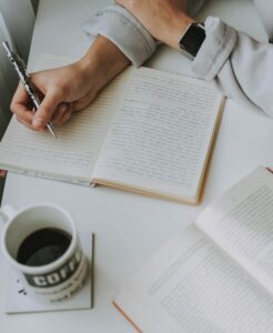 A person writing in a notebook with a cup of coffee and an open book on a white table.